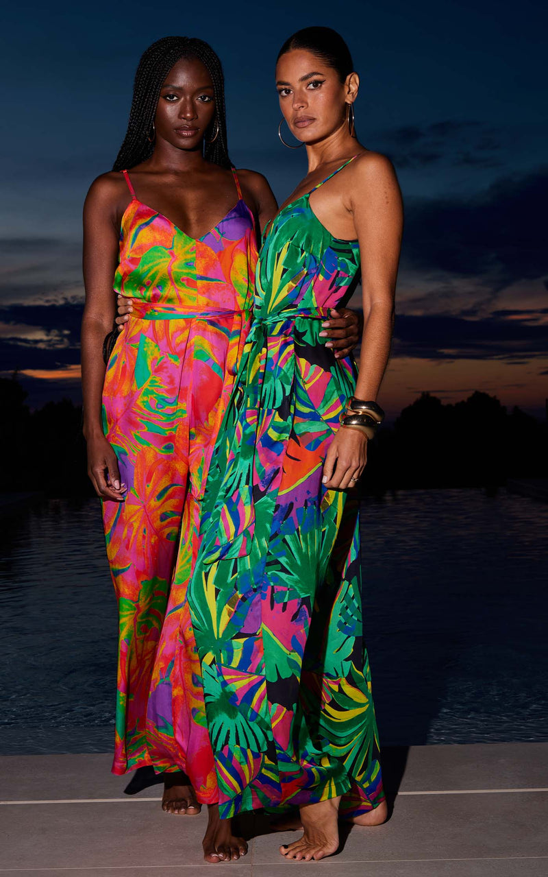 Two female models face forward in front of a pool. One wears a Dancing Leopard green, tropical print jumpsuit and the other wears a pink tropical print jumpsuit.