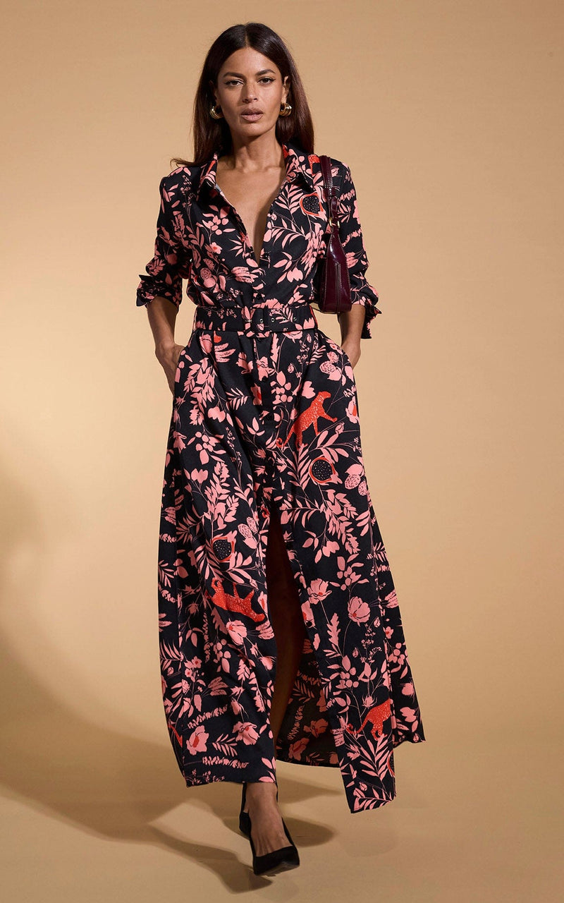 A female model faces forwards with her hands in her pockets. She wears a Dancing Leopard maxi shirt dress. The dress is black with a orange flower print.