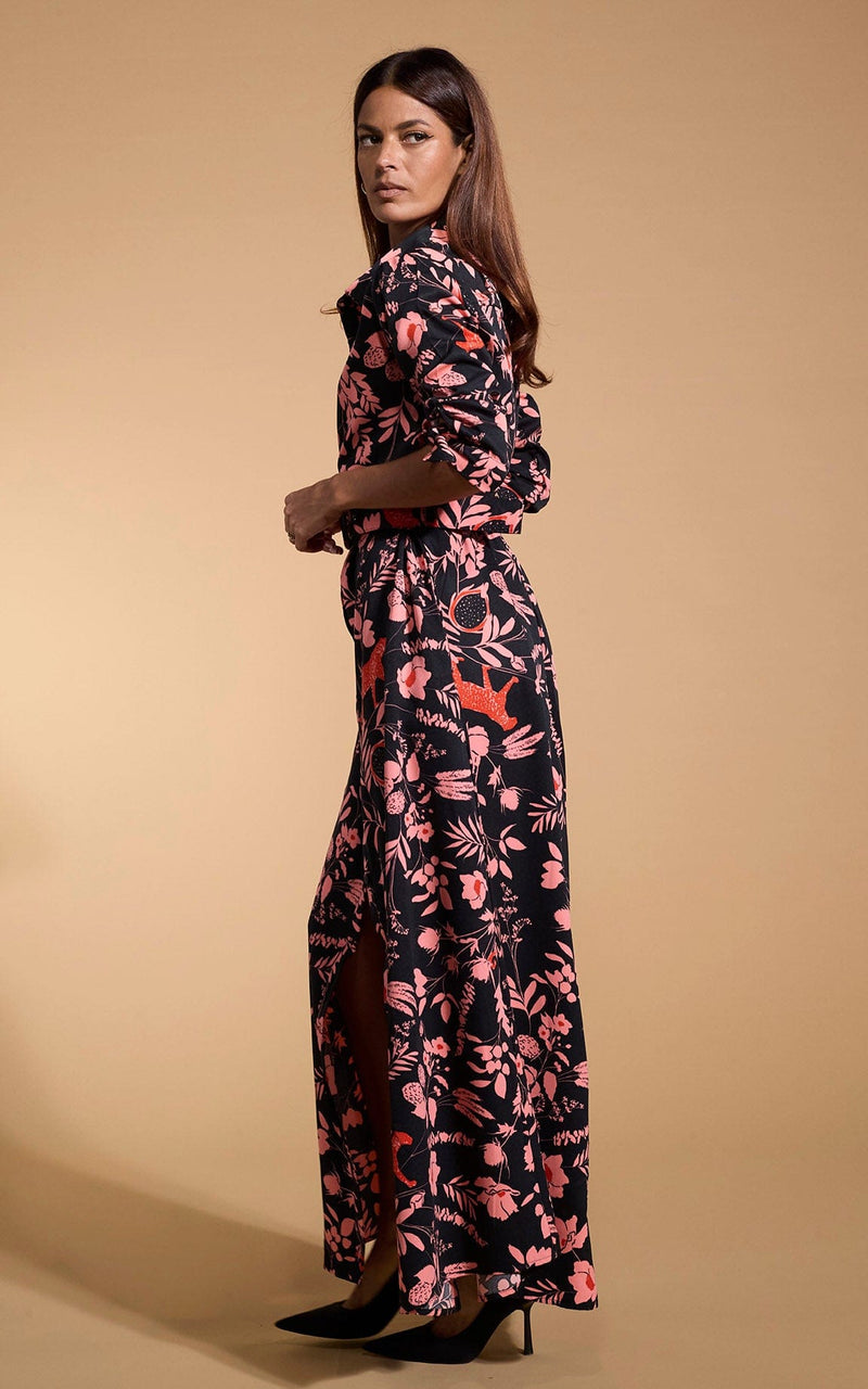 A female model faces side on. She wears a Dancing Leopard maxi shirt dress. The dress is black with a orange flower print.