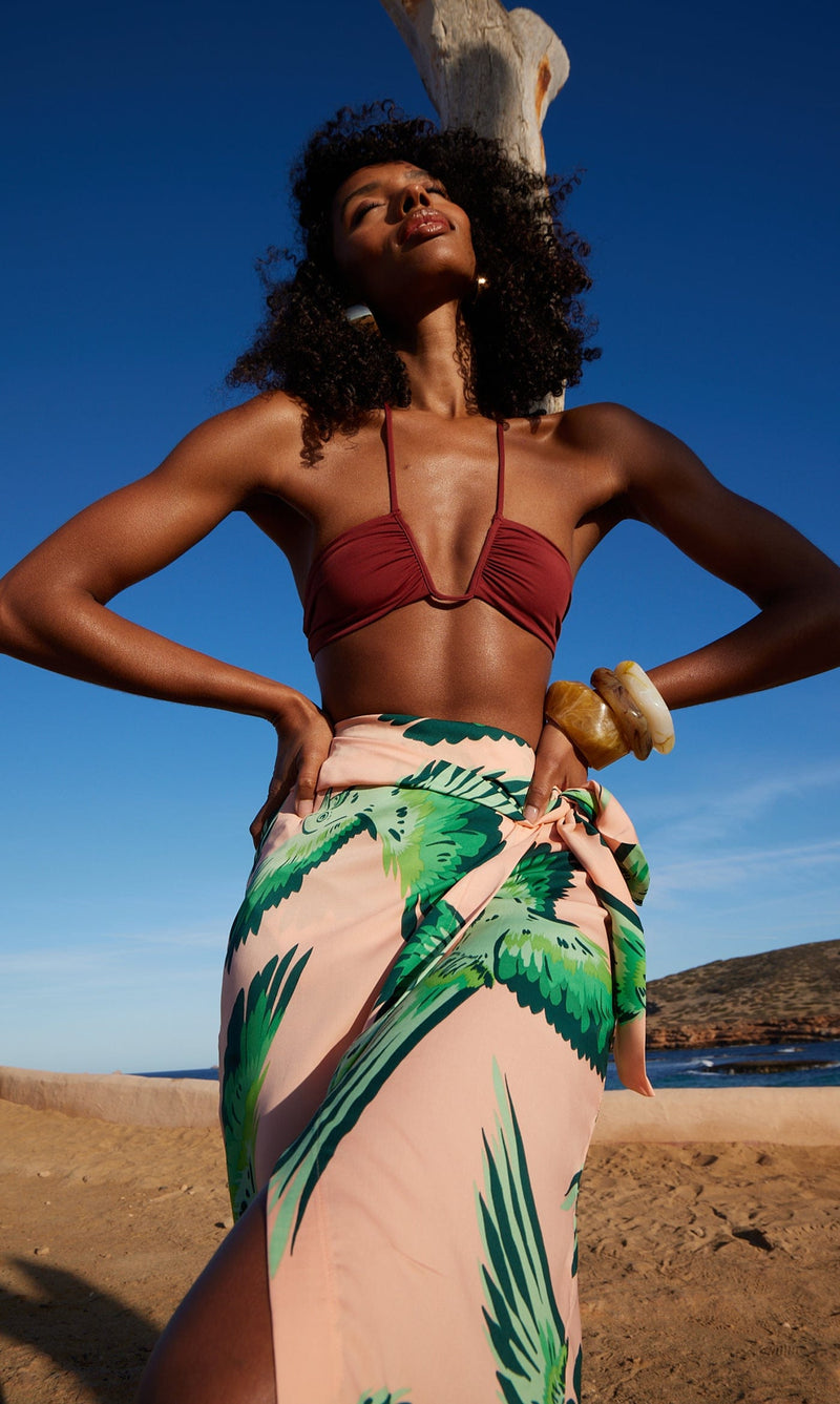 A female model faces forwards in front of the sea. She wears a Dancing Leopard peach, parrot print skirt with a maroon bikini top.