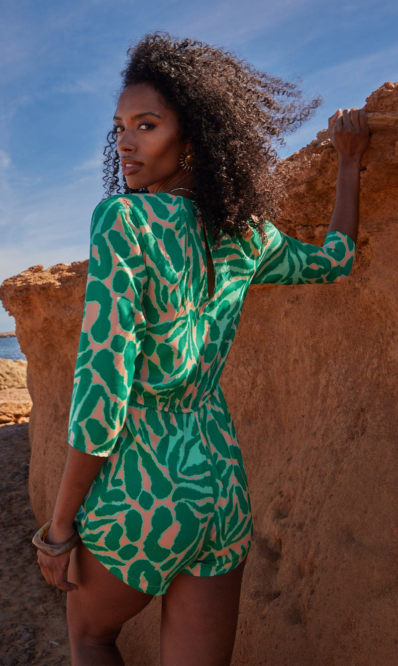 A female model faces backwards, leaning against a rock. She wears a Dancing Leopard green and peach, zebra print playsuit.