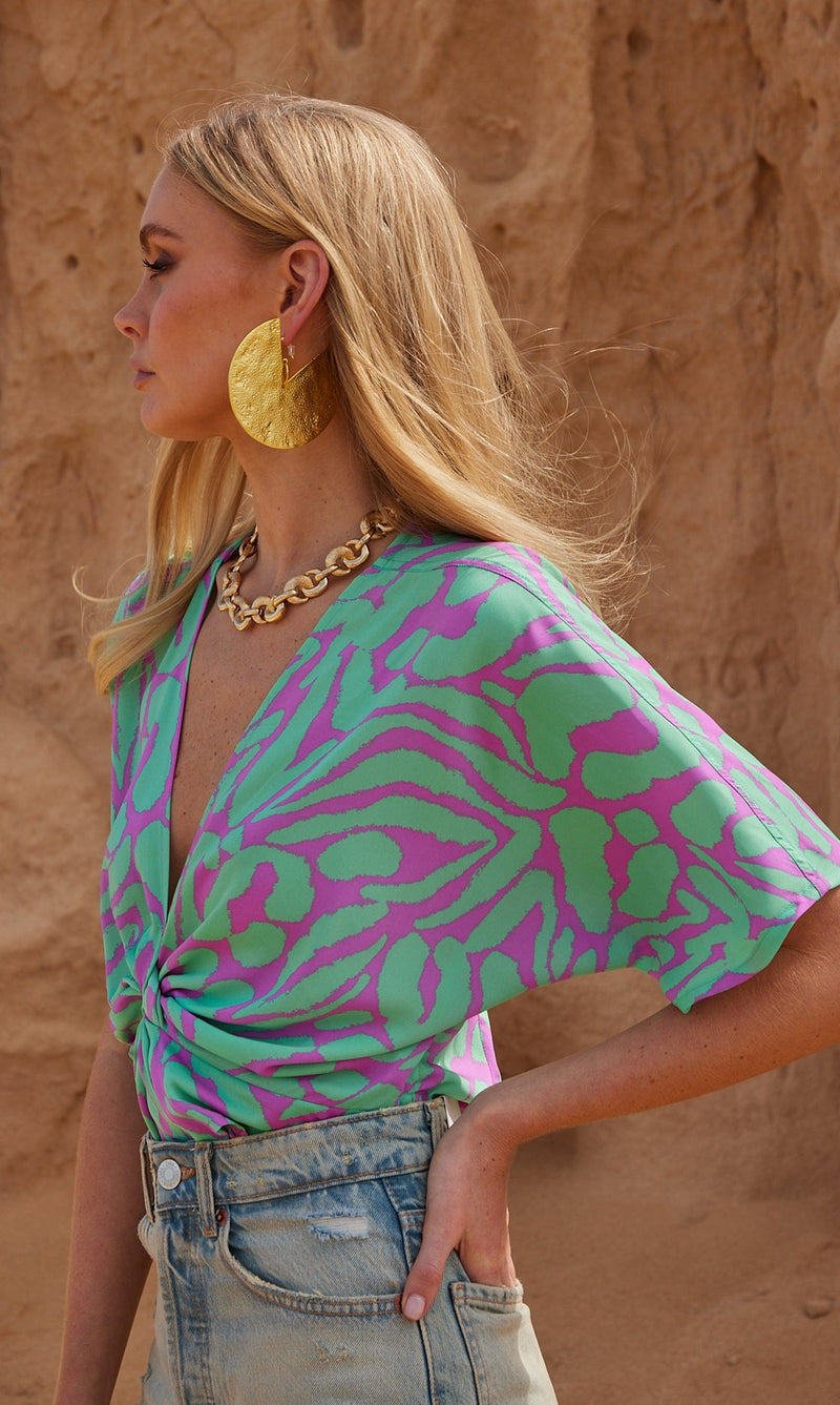 A female model faces forwards. She wears a Dancing Leopard purple and mint green, zebra print top with denim shorts.