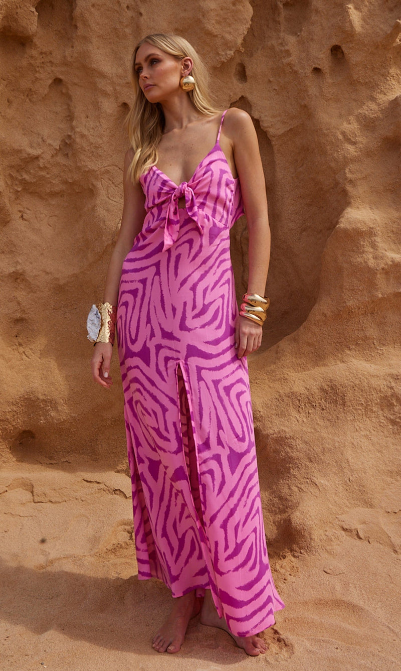 A female model faces forwards in front of a sea cliff. She wears a Dancing Leopard pink, marble print maxi dress.