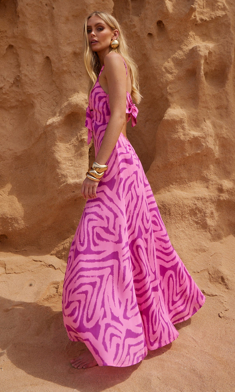 A female model faces forwards in front of a sea cliff. She wears a Dancing Leopard pink, marble print maxi dress.