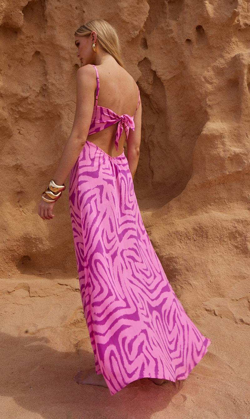 A female model faces backwards in front of a sea cliff. She wears a Dancing Leopard pink, marble print maxi dress.