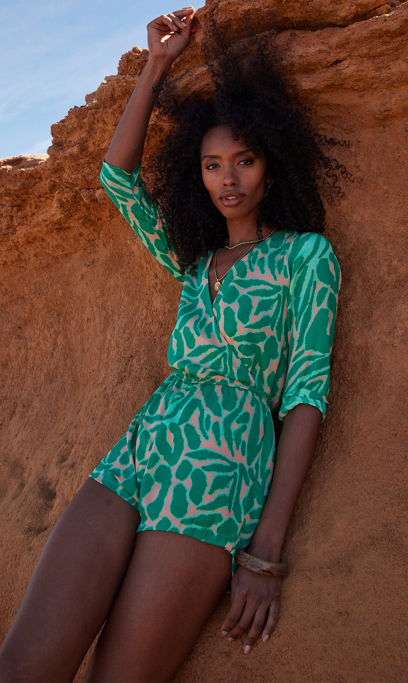 A female model faces forwards, leaning against a rock. She wears a Dancing Leopard green and peach, zebra print playsuit.