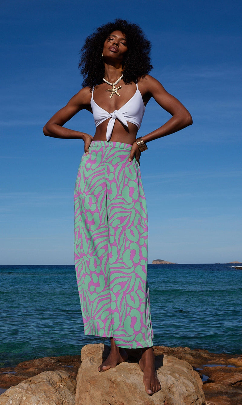 A female model faces forwards standing on a rock in front of the sea. She wears Dancing Leopard purple and mint, zebra printed trousers with a white bikini top.