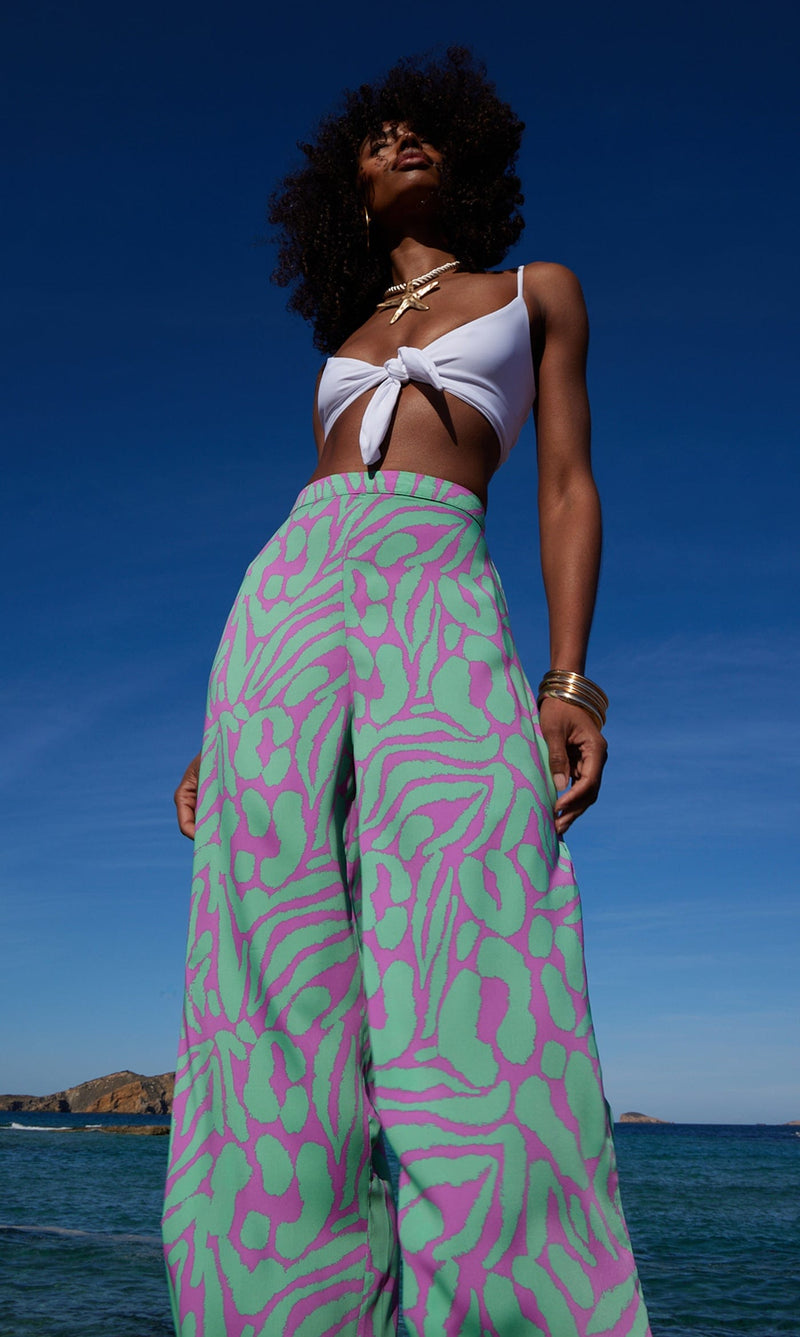 A female model faces forwards in front of the sea. She wears Dancing Leopard purple and mint, zebra printed trousers with a white bikini top.