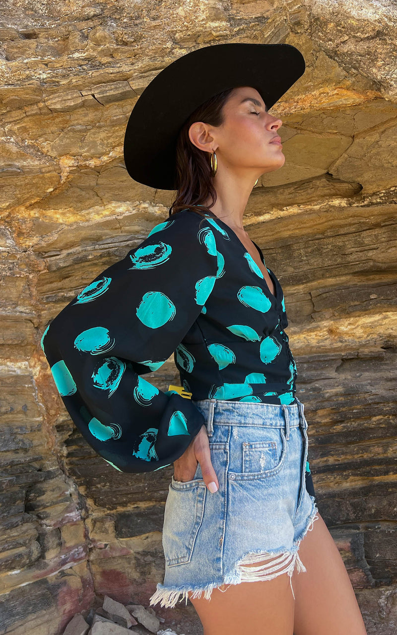 A female model faces forward. She wears a Dancing Leopard black with green dots long sleeved top. The top is paired with denim shorts and a black hat.