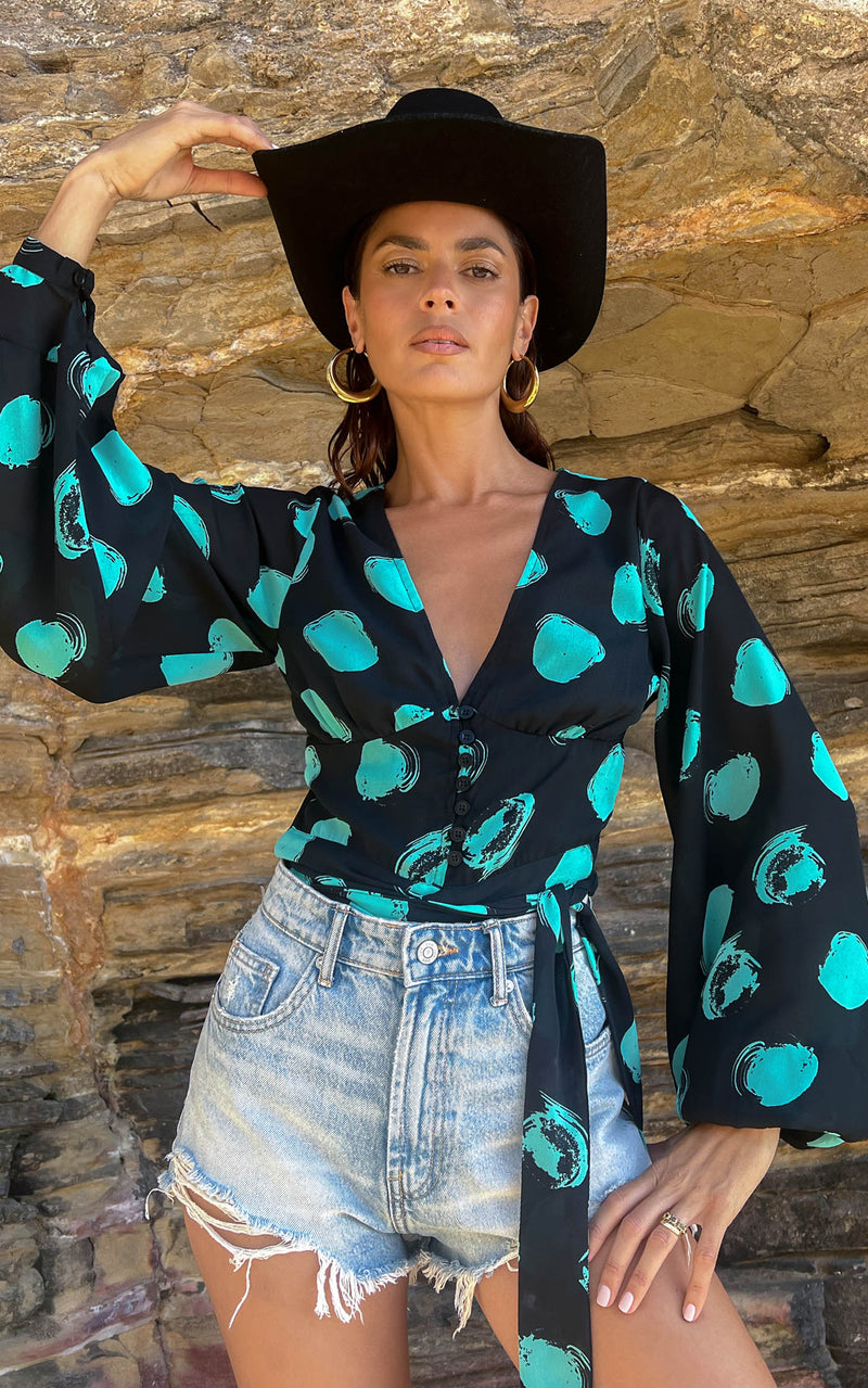 A female model faces forward. She wears a Dancing Leopard black with green dots long sleeved top. The top is paired with denim shorts and a black hat.