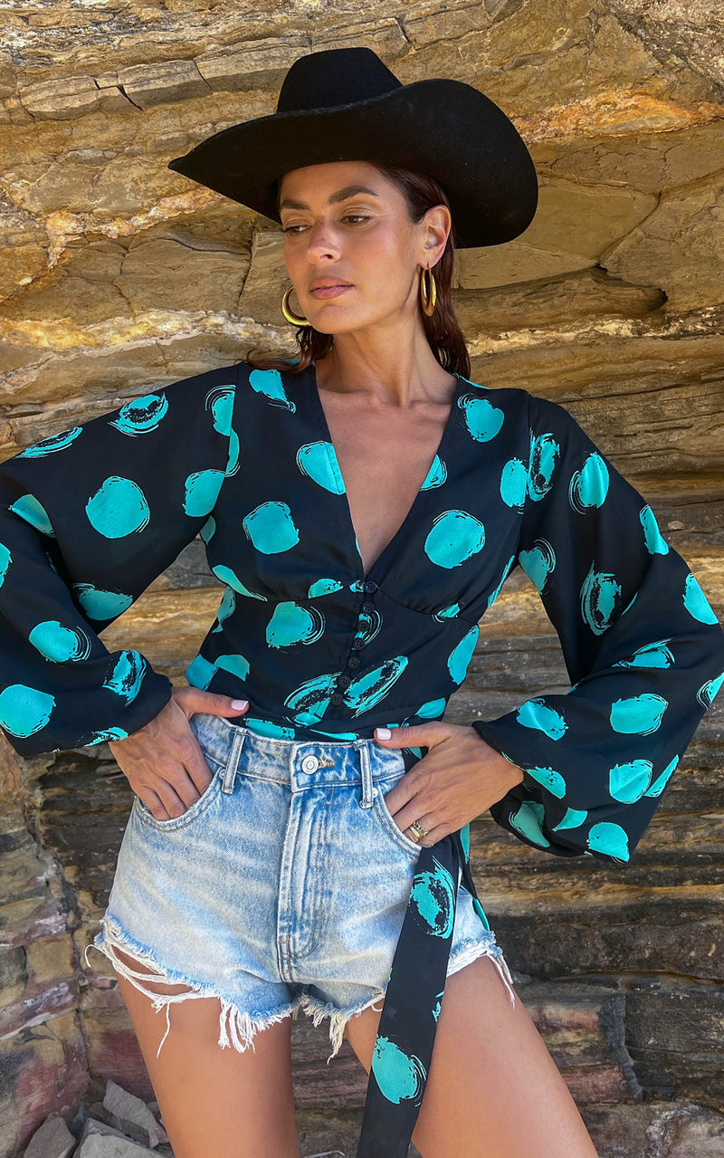 A female model faces forward. She wears a Dancing Leopard black with green dots long sleeved top. The top is paired with denim shorts and a black hat.