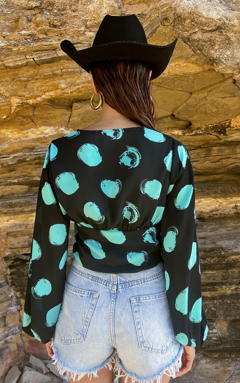 A female model faces backwards. She wears a Dancing Leopard black with green dots long sleeved top. The top is paired with denim shorts and a black hat.