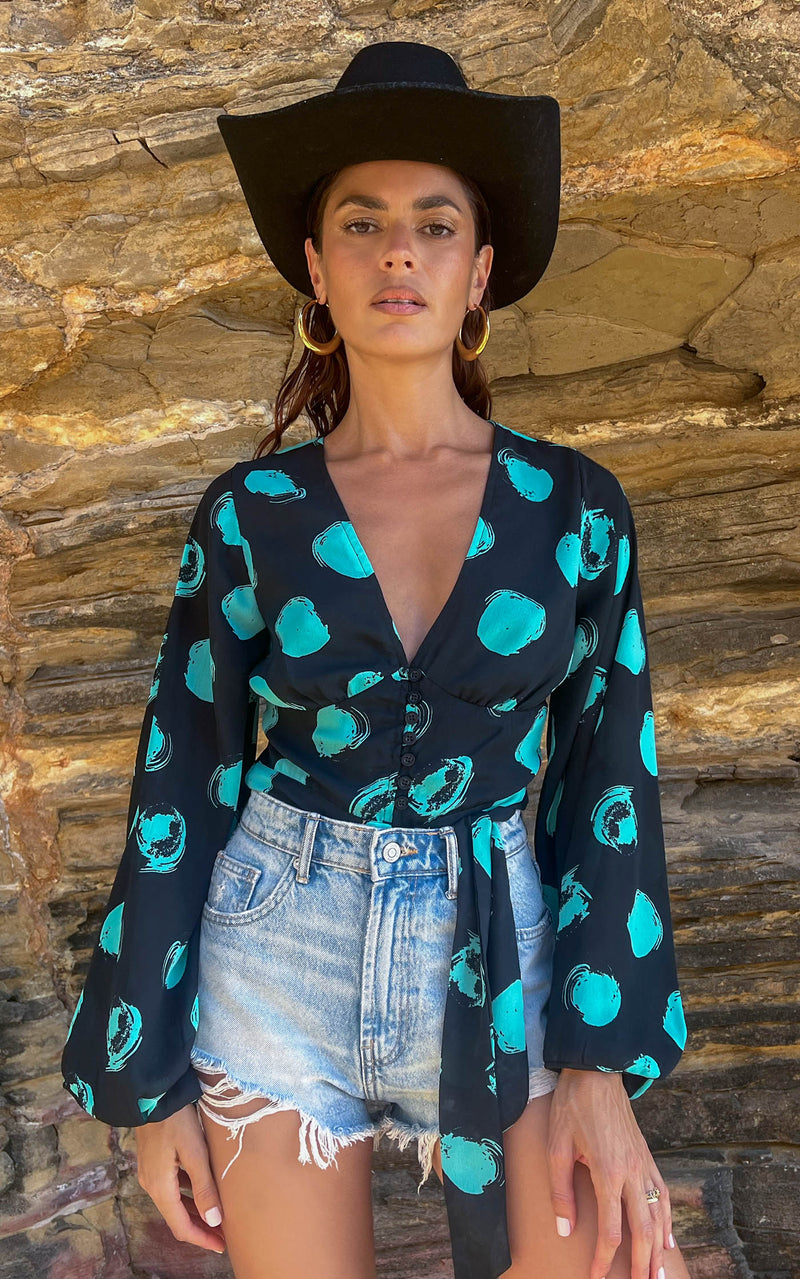 A female model faces forward. She wears a Dancing Leopard black with green dots long sleeved top. The top is paired with denim shorts and a black hat.