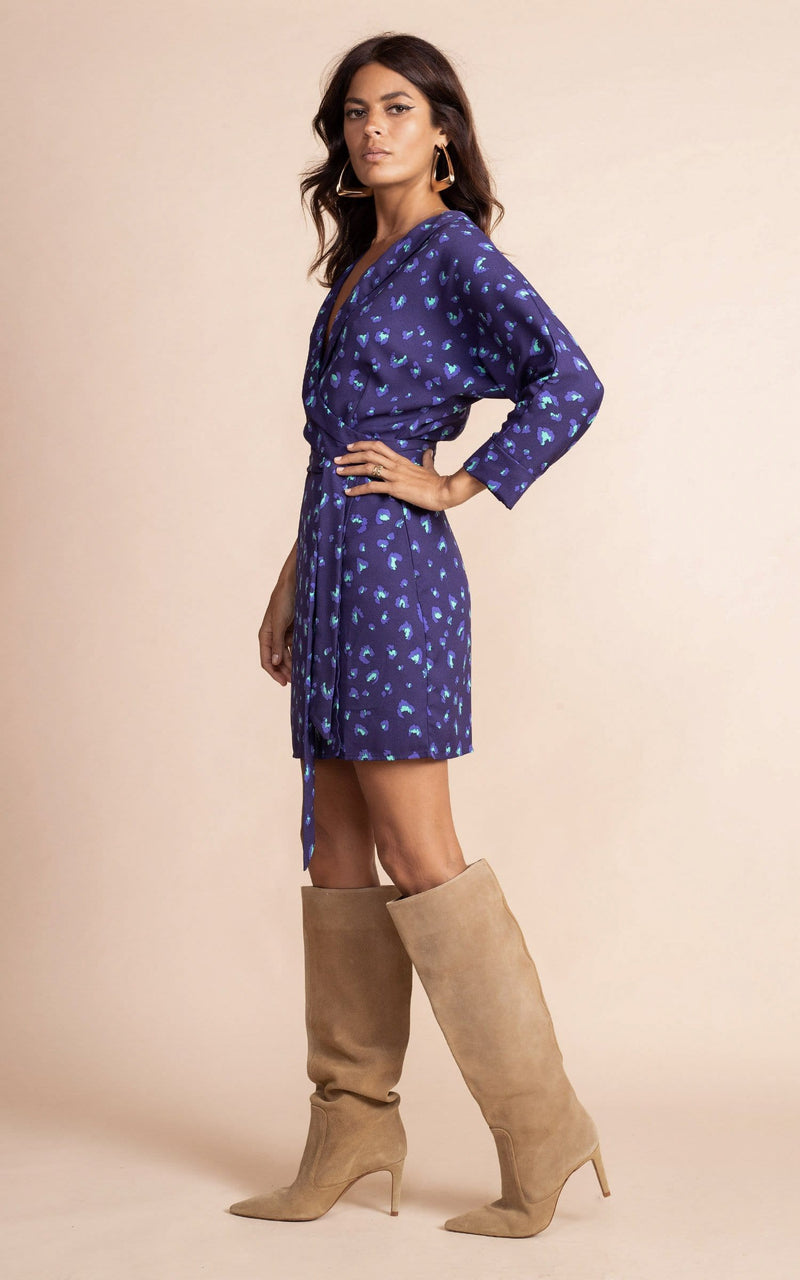 Dancing Leopard model standing sideways to the camera and wearing navy blue and purple mini wrap dress in leopard print, with tanned knee high boots